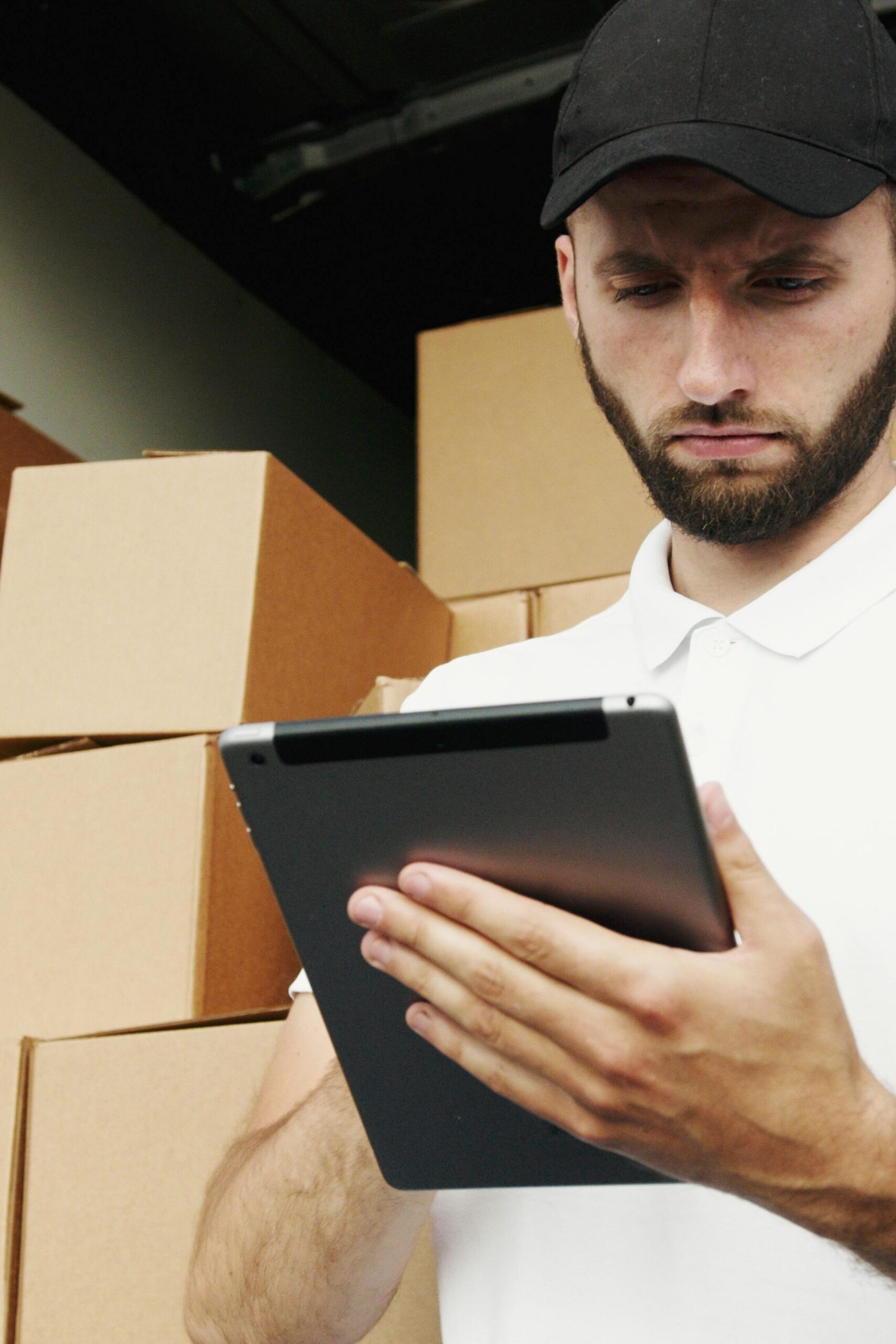 pexels-photo-5025643-5025643 Delivery worker using a tablet to manage shipments with stacked boxes in the background.